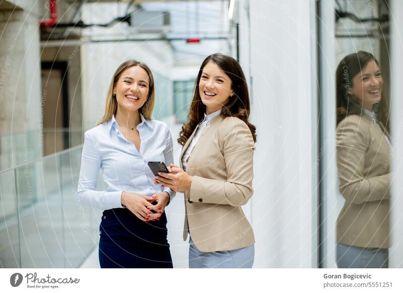 Two young business women with mobile phone in the office hallway adult businessman businesspeople businesswoman businesswomen caucasian ethnicity communications