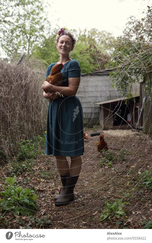Portrait of young woman with flower wreath in garden with brown chicken in her arms - in rubber boots and dress in chicken run Woman Young woman Dress Garden