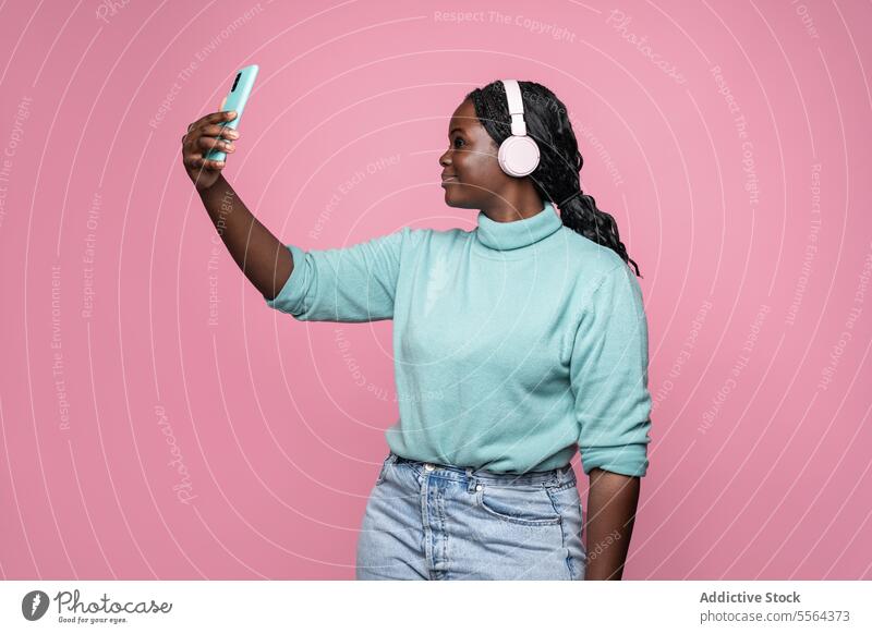 African woman enjoying music against a pink background headphone smartphone teal sweater listen audio entertainment profile leisure african braids device hand