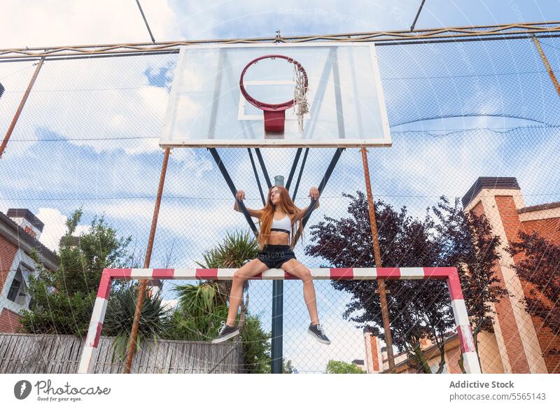 Redhead woman hanging on basketball ring in city setting redhead sky outdoors strength agility sports bra black shorts athletic determination urban playground