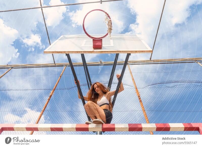 Redhead woman hanging on basketball ring in city setting redhead sky outdoors strength agility sports bra black shorts athletic determination urban playground