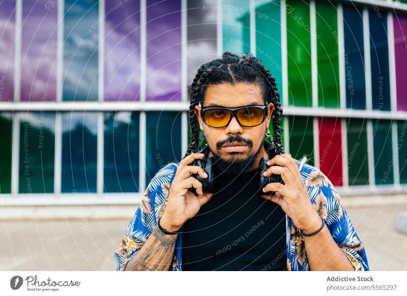 Latin man adjusting headphones against a colorful building windows. latin braids holding sitting sunglasses music background diversity outdoors shirt look front