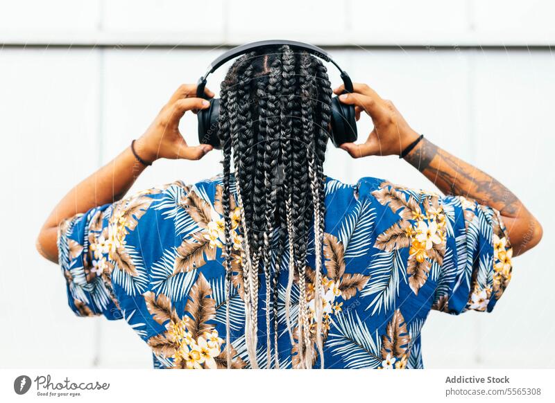 Latin man listening music with his black headphones from behind against a white background. braids tattoos floral blue shirt rear view adjusting hands detail