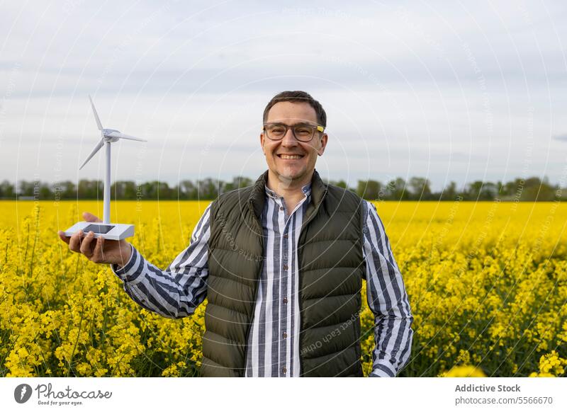 Mature man with glasses showcasing a wind turbine model in bloom-filled meadow. field flower yellow present smile nature stand showcase green spring renewable