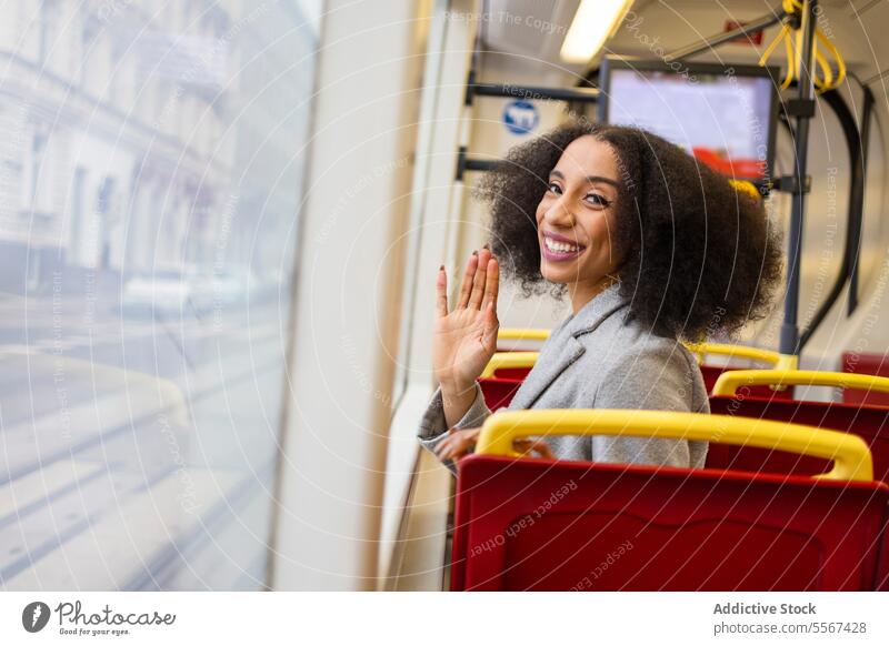 Young afro woman waving hello on bus curly hair window reflection city transportation joy seat red commute public travel urban journey coat grey comfort transit