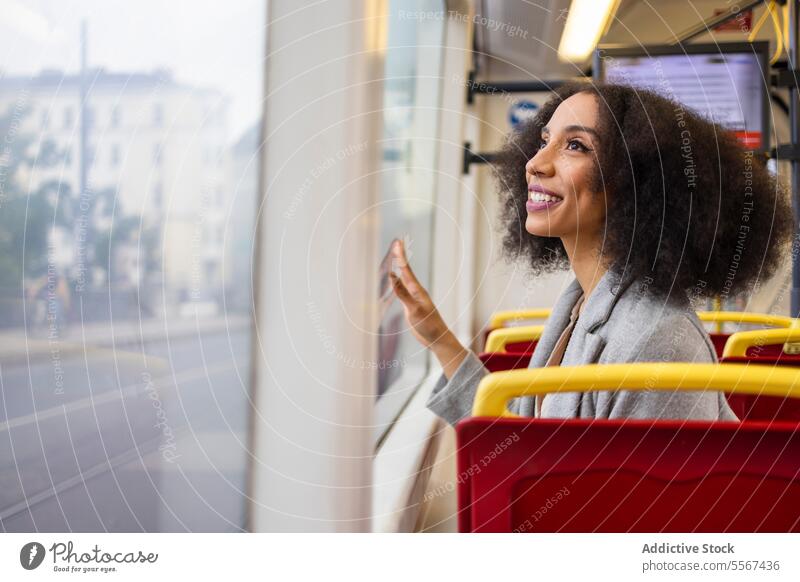 Happy afro woman looking though window while sitting on bus seat curly hair reflection city transportation joy admire red commute public travel urban journey