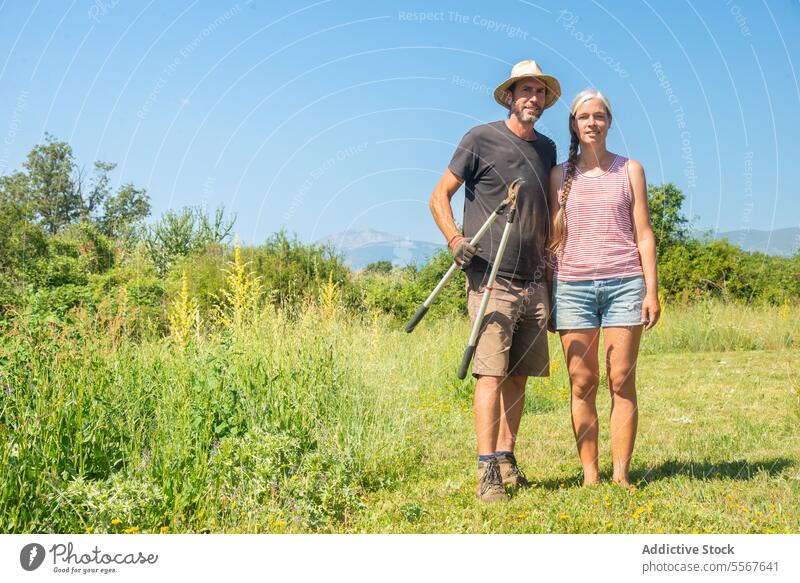 Mature couple with tools standing on open field Man woman hat straw gardening striped tank top mountains background outdoors nature sunny rural landscape