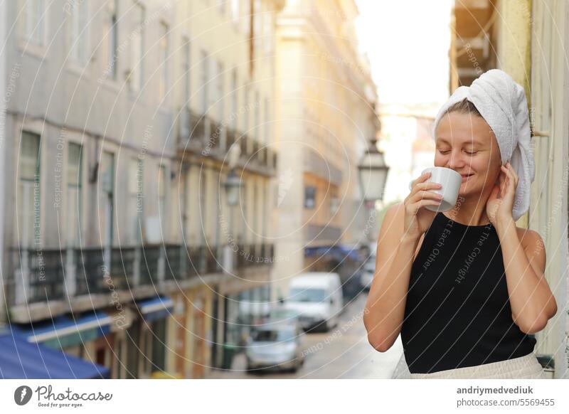 Young beautiful woman in a towel on head is drinking coffee or tea and smiling cute standing on balcony on ueropean street background. lifestyle morning. Concept of domestic or traveling lifestyle.