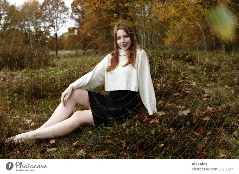 Young woman in white wool sweater and black skirt sits barefoot in a northern German heath landscape in the fall in the grass between heather and smiles Woman