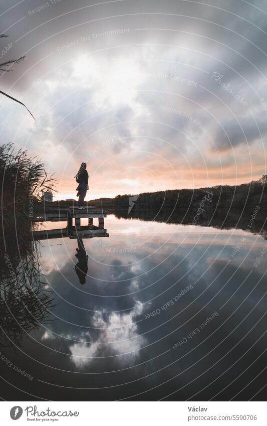 Lady aged 20-25 standing on a wooden pier by a pond watching the sunset over the town of Oostende, western Belgium. Dramatic red-orange sky female exploration