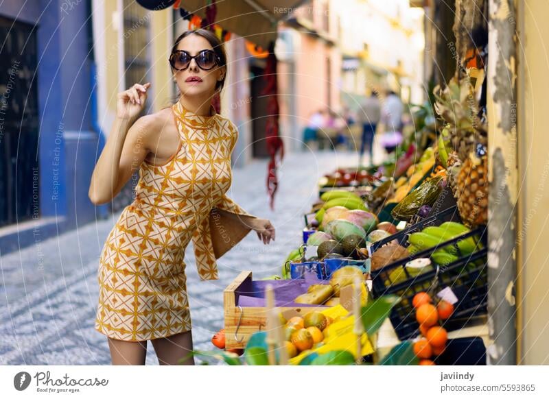 Stylish young woman standing by assorted fruit shop in street style positive trendy fashion town stall smile eyeglasses makeup lean forward content fresh