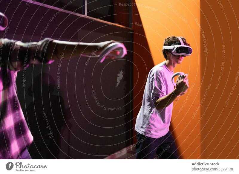 Side view of two friends stand in a gaming area fully immersed in a virtual reality world wearing VR headsets and holding motion controllers in neon light room
