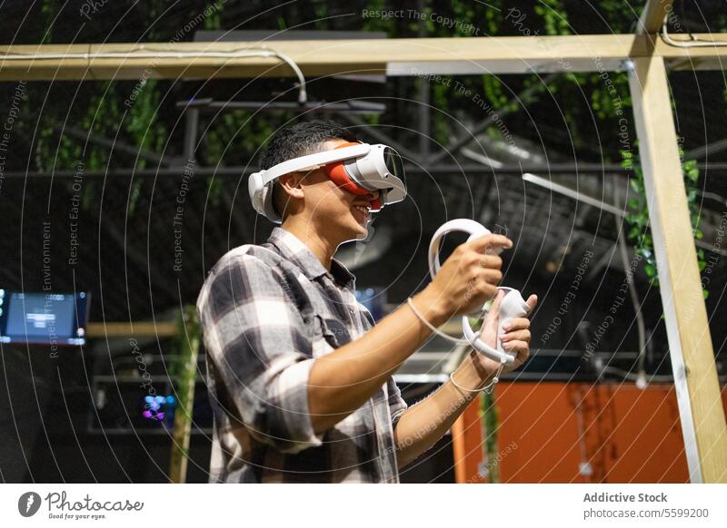 Side view of young excited man is engaged in a virtual reality game wearing a VR headset and using controllers technology vr goggles gadget entertainment