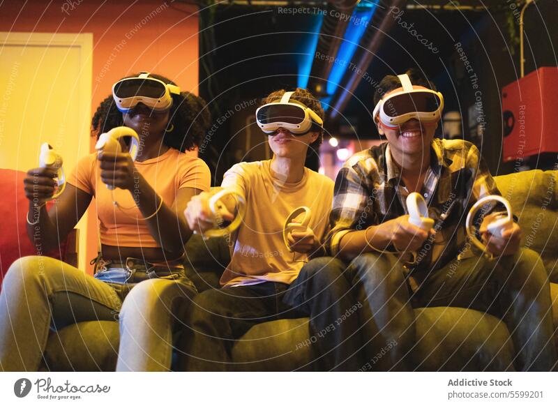 Multiethnic friend engrossed in a virtual reality game wearing VR headsets and holding controllers sitting on a couch in a dimly lit room vr friends technology