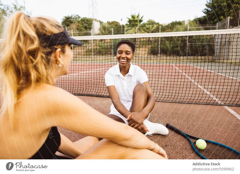 Two young beautiful women having a conversation sitting on a tennis court next to the net. Two amateurs tennis players having a break during a tennis match.