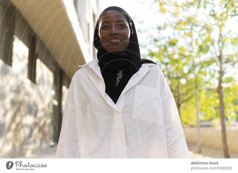 From below happy African American young confident businesswoman in black headscarf looking away while standing against building and trees in city hijab