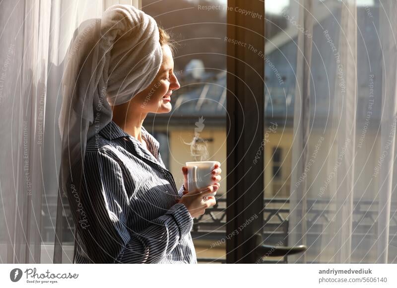 Beautiful young woman with bath towel on head holds a white cup of hot coffee or tea at home by the window. Smiling girl after shower is enjoying aroma drinks and have breakfast in the morning