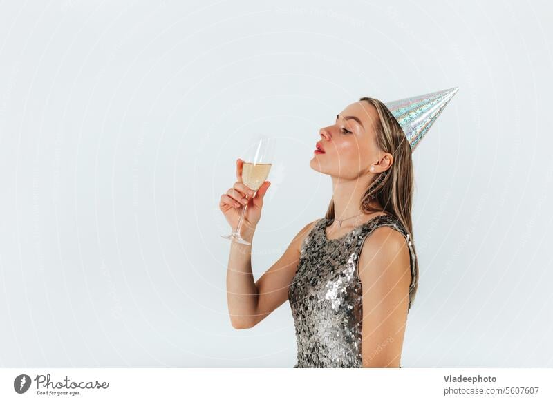Young caucasian blonde woman with wine glass wearing silver glitter dress and party hat celebrating on white background champagne party cap shiny holiday
