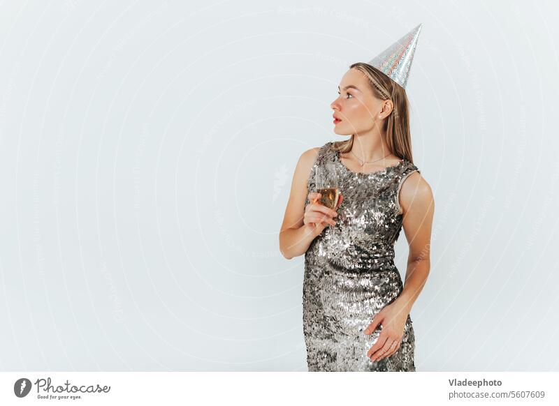 Young caucasian blonde woman with wine glass wearing silver glitter dress and party hat celebrating on white background. champagne party cap shiny holiday