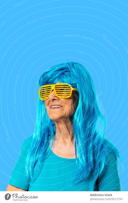 Smiling elderly woman in sunglasses and blue wig looking away while standing on blue background portrait stripe retire pensioner funny wrinkle accessory smile