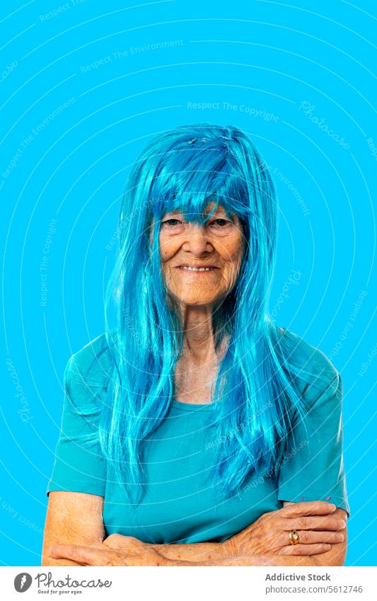 Smiling elderly lady in blue wig looking at camera while standing with crossing arms on blue background woman portrait hands crossed arms crossed appearance