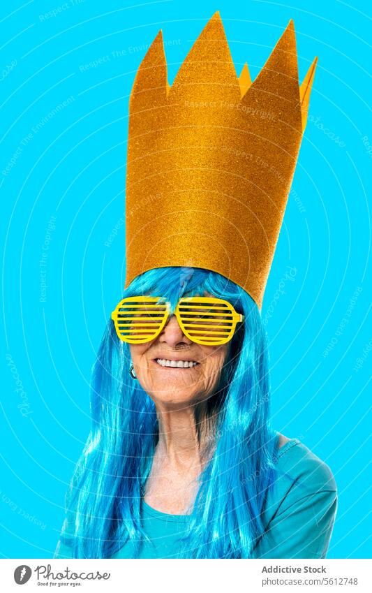 Funny aged woman wearing blue wig, golden paper crown and sunglasses while looking at camera on blue background portrait stripe retire wrinkle pensioner funny