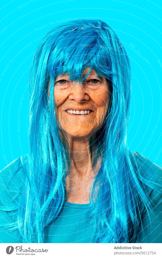 Smiling elderly lady in blue wig looking at camera while standing on blue background woman portrait appearance retire wrinkle pensioner funny smile positive