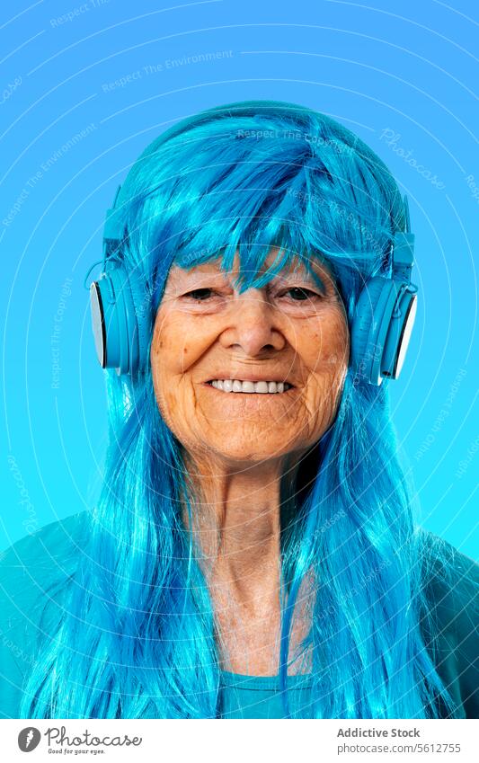Cheerful aged woman listening to music via headphones while looking at camera against blue background portrait enjoy song wig color match smile happy cheerful