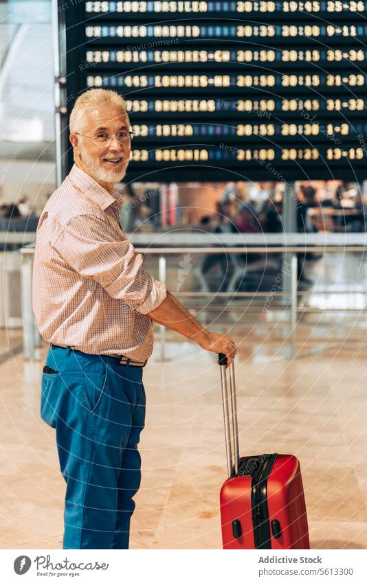 Man with luggage looking at camera at airport senior man departure board smile timetable side view suitcase vacation travel retired lifestyle holiday leisure