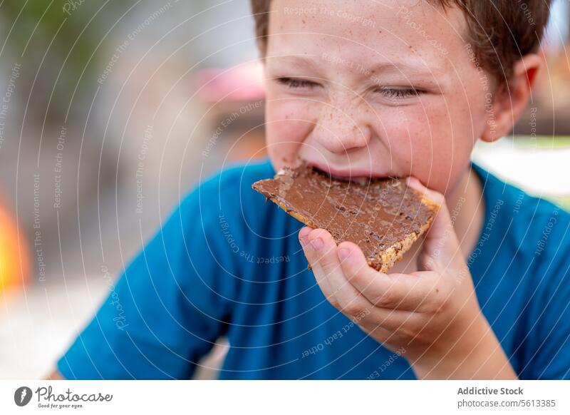 Cute hungry boy dressed in casuals with eyes closed eating sweet chocolate spread with bread for breakfast while sitting in restaurant sandwich cute expression