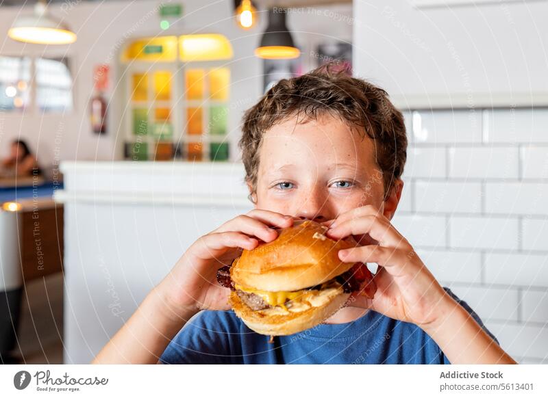 Portrait of hungry cute elementary boy eating delicious burger and looking at camera while spending leisure time in restaurant at weekend innocent meal portrait