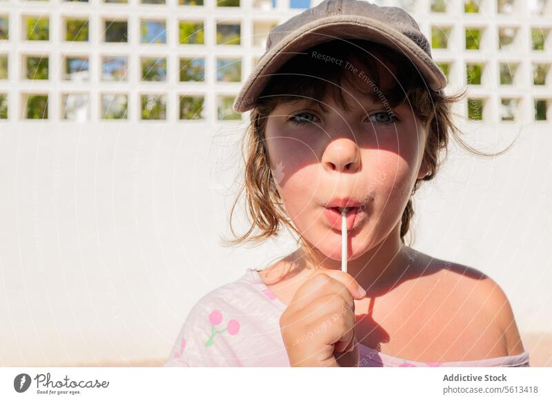 Closeup portrait of cute serious girl in casual clothes and cap eating sweet lollipop and staring at camera while standing on the street caucasian innocent cafe