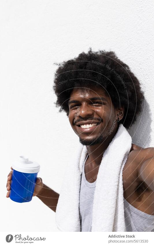 Smiling young male athlete with afro hair holding water bottle and taking selfie looking at camera while leaning on white wall after workout in summer man