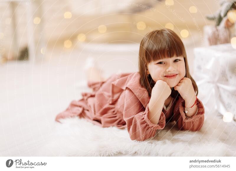 little cute child girl in pink dress lying on white furry carpet in decorated Christmas room interior and looking in camera. Beautiful bokeh. Happy family New Year, Xmas concept
