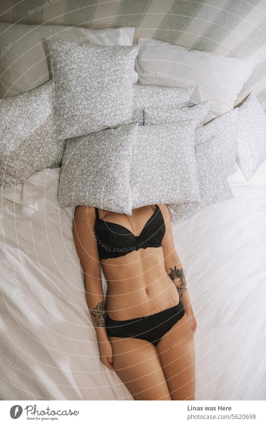 A gorgeous lingerie model in black panties and black bra is covering her face under the pillows. A sleeping inked beauty is feeling relaxed in her own skin and in her own underwear. Her sun-kissed body lies gracefully upon the bed.