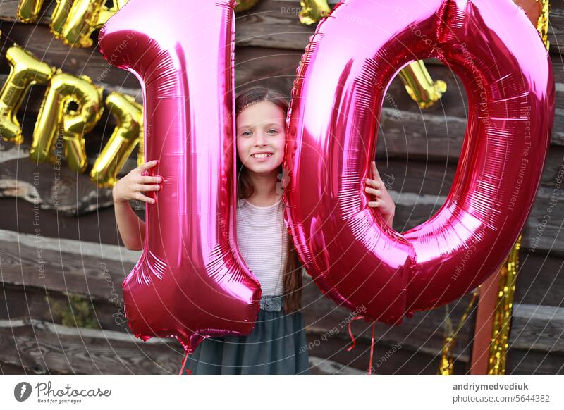 Cheerful little girl holds two big pink foil balloons, number 10 outdoors on wooden wall background with golden balloons inscription happy birthday. Ten years anniversary celebration party at home