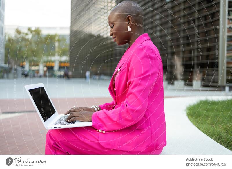 Side view of black businesswoman with shaved head in pink clothes working on laptop on street entrepreneur independent self employed browsing typing freelance