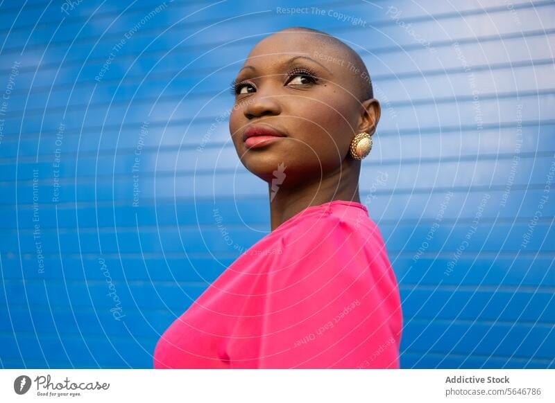 Confident black woman with shaved head in pink dress standing against blue wall outfit african american ethnic bald female style stripe trendy modern delight