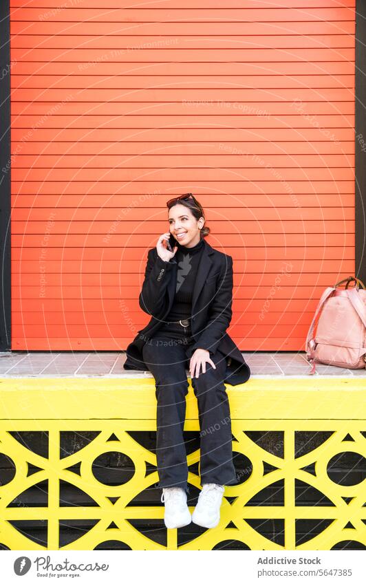 Young woman sitting on orange wall and talking on smartphone phone call speak smile happy street conversation cheerful female young mobile dress device