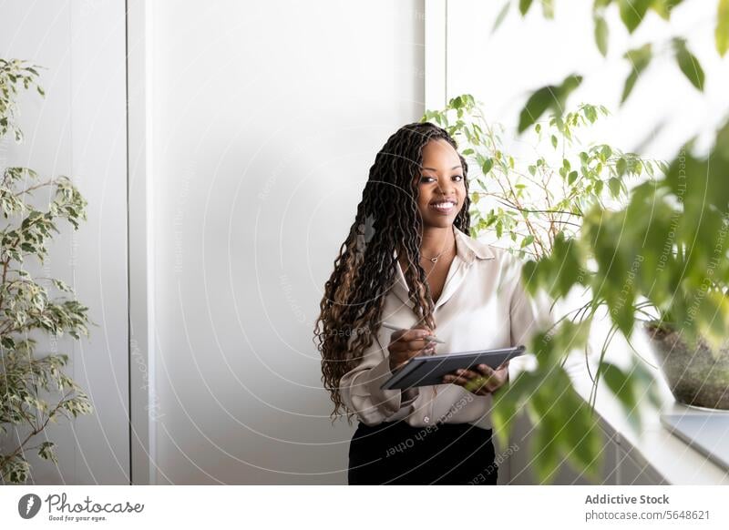 Thoughtful young African American businesswoman with faux locs hairstyle holding digital tablet and stylus while looking at camera while stating in office