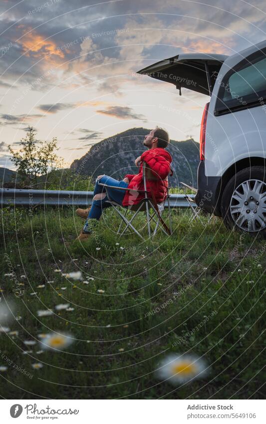 Man relaxing on chair near car in countryside at dusk Tourist Relax Chair Car Mountain Grassy Road Trip Field Traveler Countryside Sky Dusk Cloudy Jacket Cold