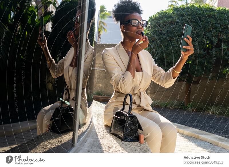 Black Entrepreneur in smart casual applying lipstick using smartphone as mirror while sitting outdoors professional mobile elegant beautiful entrepreneur glass
