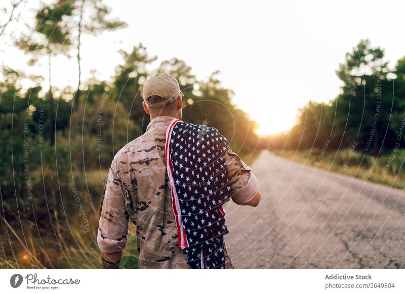 Back view of unrecognizable mature commando on road with American flag on shoulder under sky during sunset soldier american walking camouflage uniform special