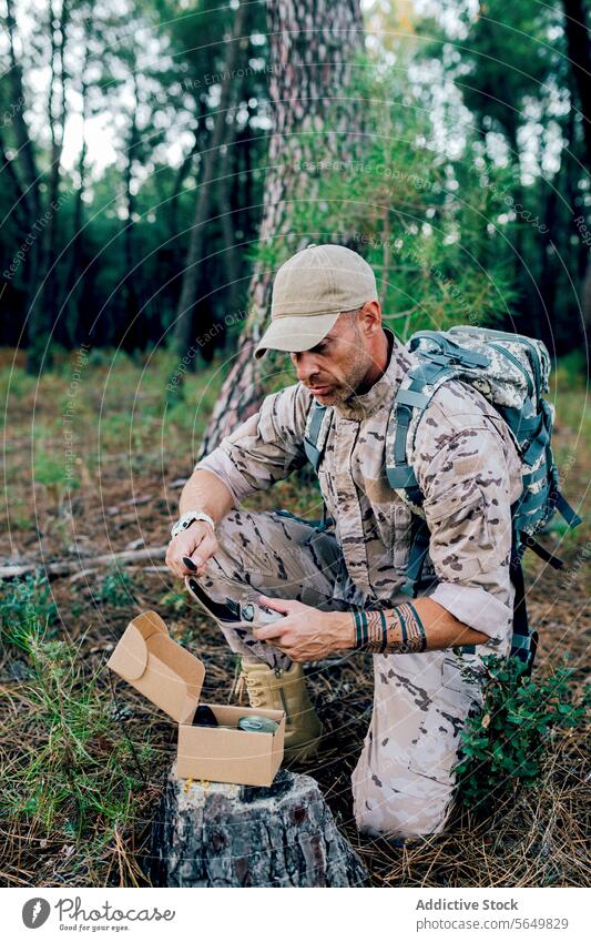 Military soldier with backpack holding cutlery pouch kneeling by box on tree man commando camouflage forest stump uniform force cap full length