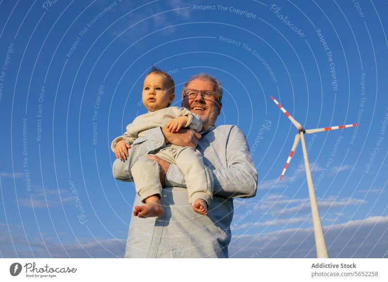 From below smiling elderly man holding a baby with a wind turbine in the background under a clear blue sky Grandfather Grandson Elderly Blue Sky grandchild