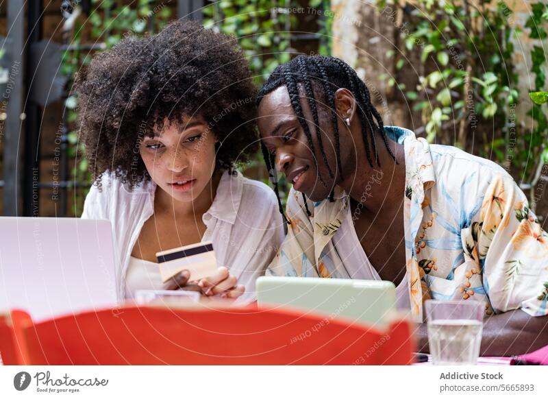 Two friends are engaged in online shopping with a woman holding a credit card and a man closely watching the laptop screen Friends black curly hair braids