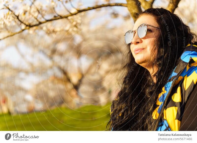 Woman in sunglasses with mirrored lenses with long dark hair in blooming spring garden in public park on sunny spring day woman cherry blossom flower tree sit