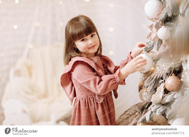 Little adorable girl in soft pink dress is hanging white shiny ball on Christmas tree with light. Child decorating Xmas tree in beautiful living room. Happy family holiday New Years concept.