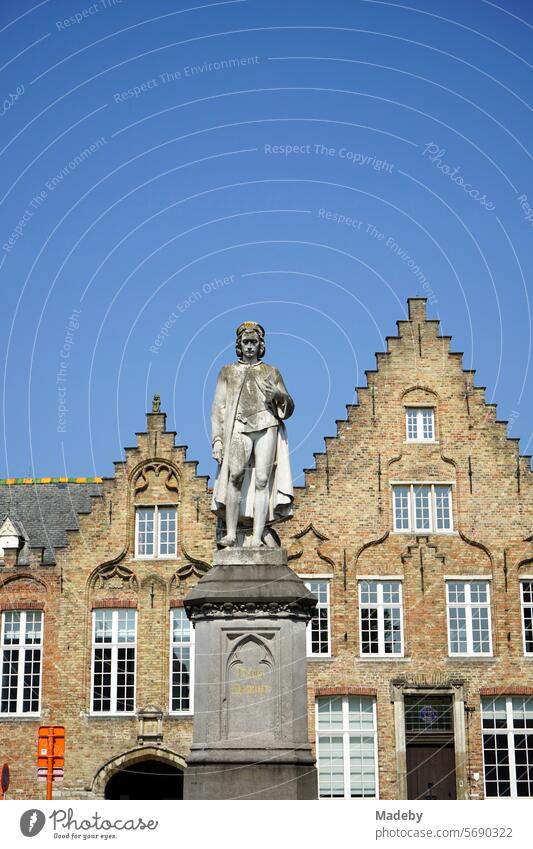 Statue of Hans Memling on the Woensdagmarkt with a beautiful old brick stepped gable against a blue sky in the sunshine in the alleys of the old town of Bruges in West Flanders in Belgium