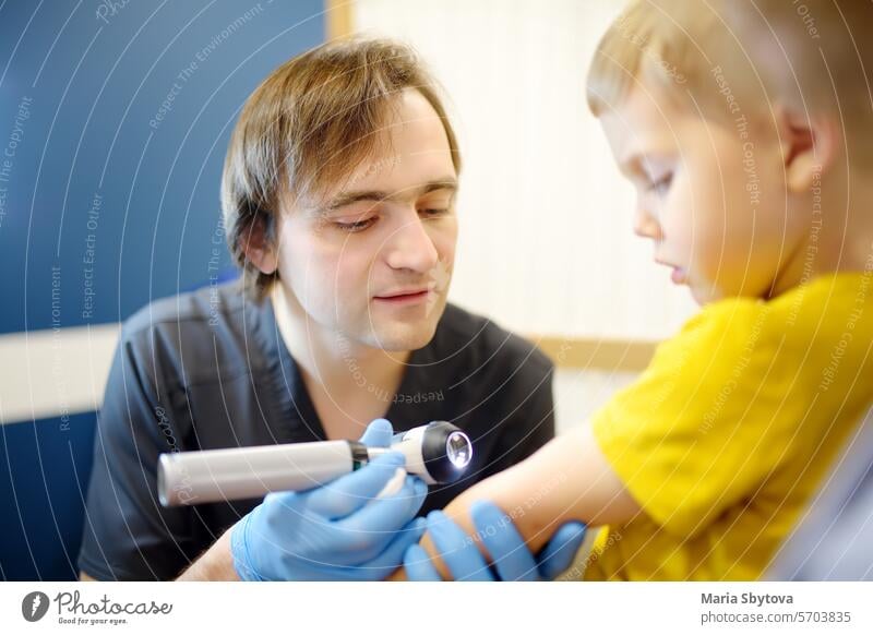 A caring doctor checks moles on the skin of a small child. A dermatologist doctor looks at a rash on the inner bend of a boy's elbow using a dermatoscope.